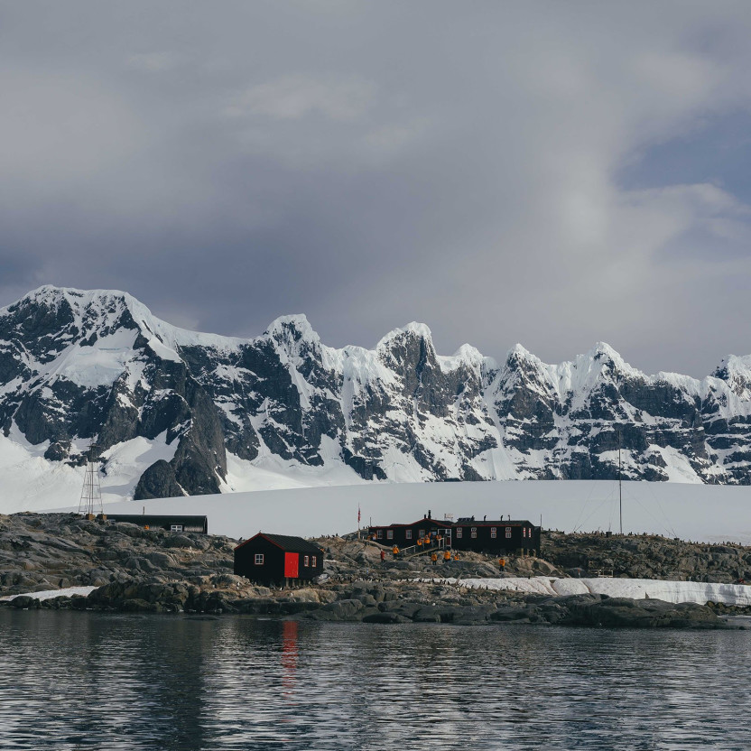 Water-level view of Port Lockroy captured from a zodiac boat. Building of the station stand out due to their red roof and guests in yellow parkas can be seen entering the post office and gift shop building.