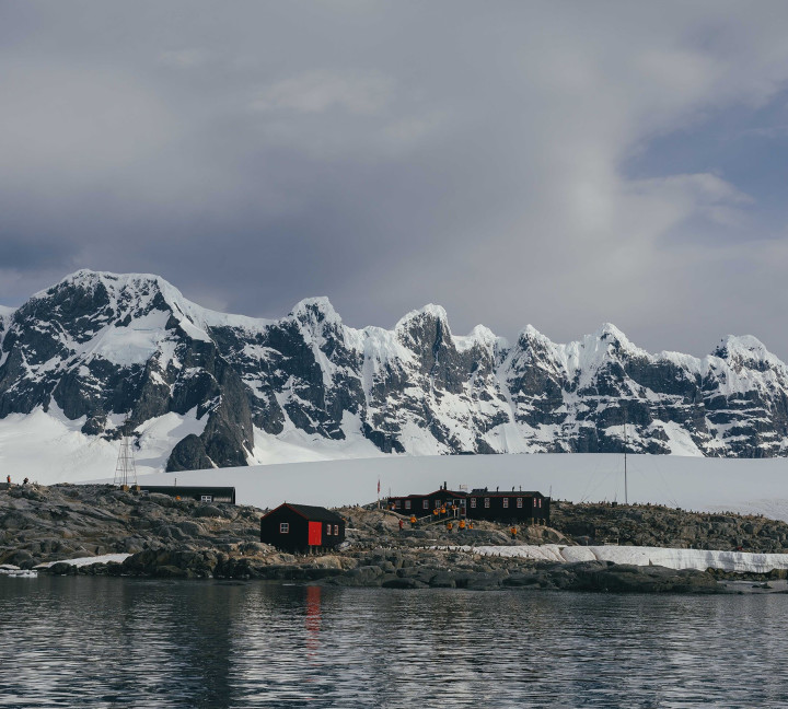 Water-level view of Port Lockroy captured from a zodiac boat. Building of the station stand out due to their red roof and guests in yellow parkas can be seen entering the post office and gift shop building.