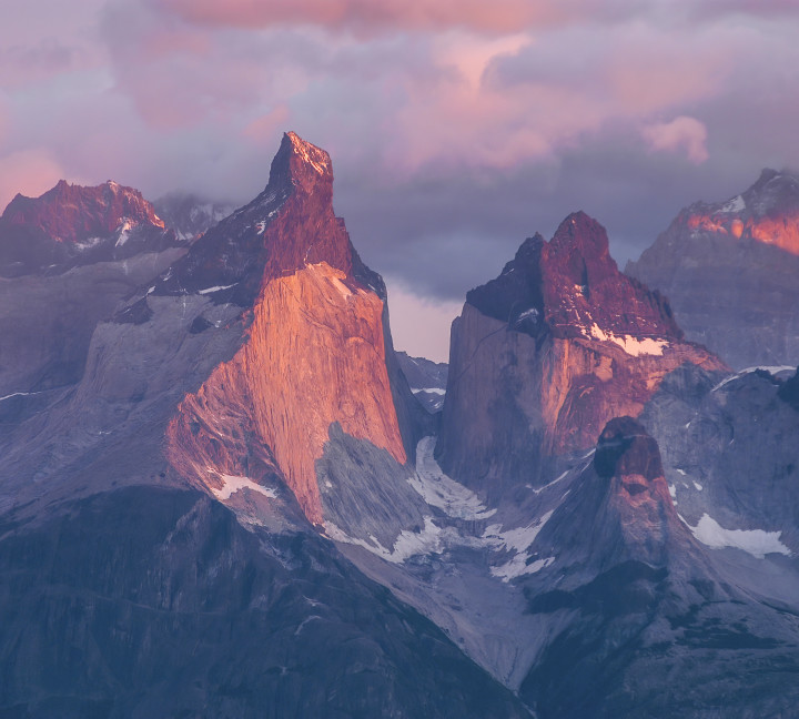 Pink and purple sunlight is cast over the spiky mountain peaks of Cuernos del Paine at Torres del Paine National Park, Chile.