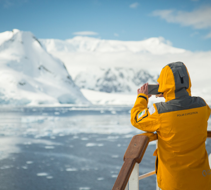 A guest in a yellow parka stands on one of the top decks of their vessel with a smartphone camera in hand.
