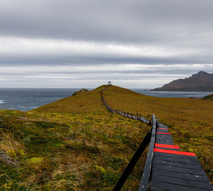 The image shows a long boardwalk stairway with the Cape Horn Monument at the end. The monument pays homage to sailors that perished attempting to round Cape Horn and depicts an albatross in flight.