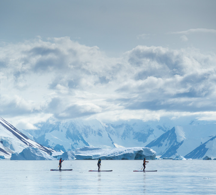 Guests go standup paddleboarding n the Antarctica Peninsula. Snow covered mountains are seen in the distance.