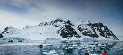 Guests in yellow parkas carefully watch an orca spyhopping as it tries to get better look at a seal resting on an ice flow.