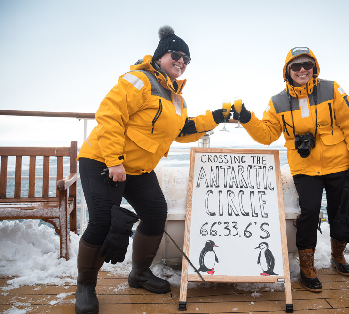 Two guests in yellow parkas pose for a photo next to a board that includes the coordinates of their ship, celebrating officially crossing the Antarctic Circle.