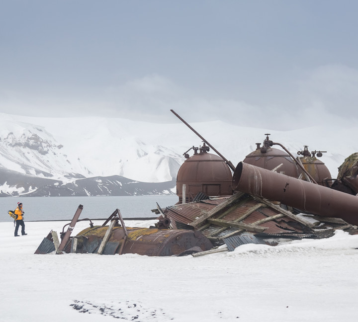 A guest in a yellow parka looks at the remnants of old and rusted equipment used to render fat from whale and seal blubber.