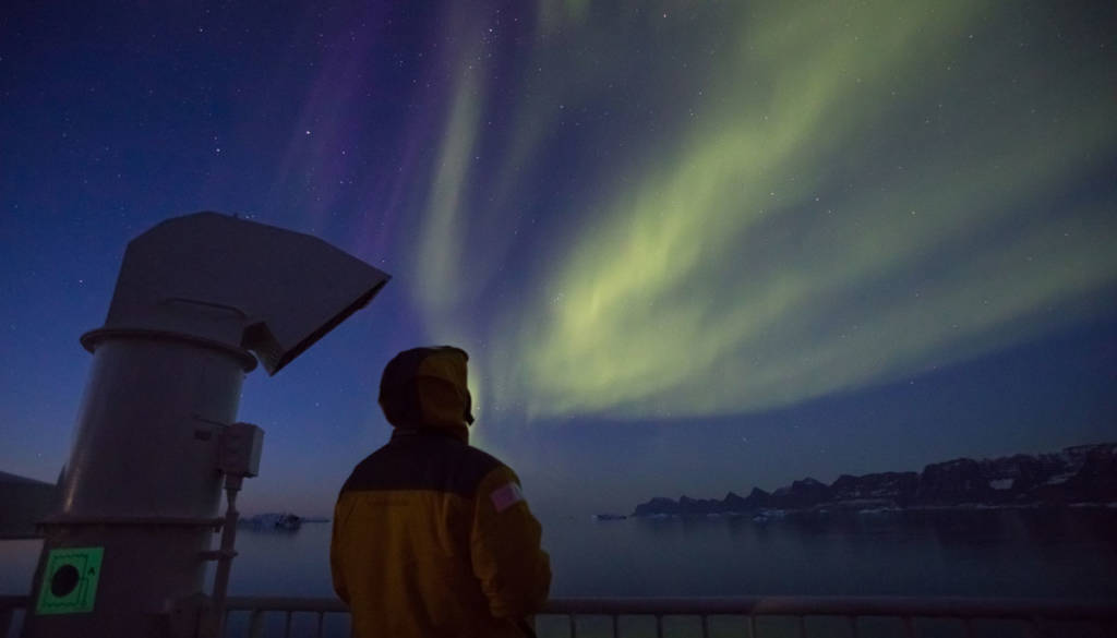 Man in a yellow jacket looks up at the streaks of Northern Lights.