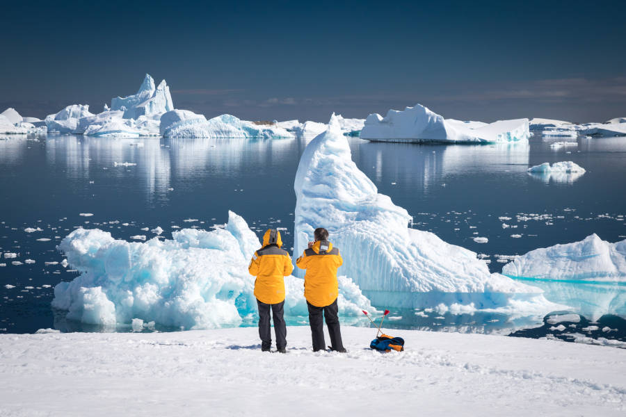 Two guests in yellow parkas admire and photograph rugged icebers off the waters of Port Charcot during a sunny afternoon in Antarctica.