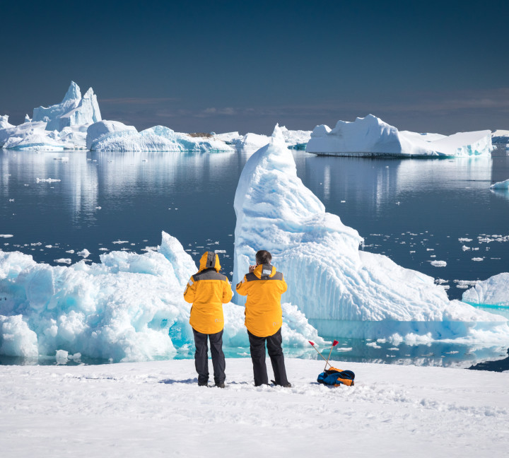 Two guests in yellow parkas admire and photograph rugged icebers off the waters of Port Charcot during a sunny afternoon in Antarctica.