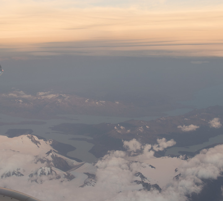View from the window of an aircraft featuring its wing and a small cloud cover over the partially ice-covered peaks near Punta Arenas, Chile.